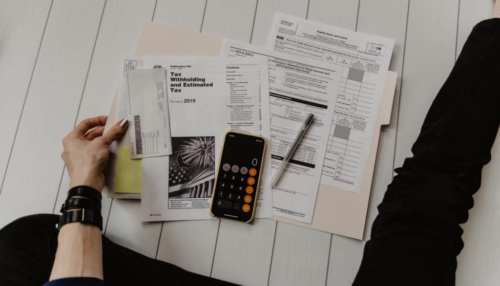 The image shows a person sitting on the floor, surrounded by various tax forms, a calculator, a pen, and envelopes.