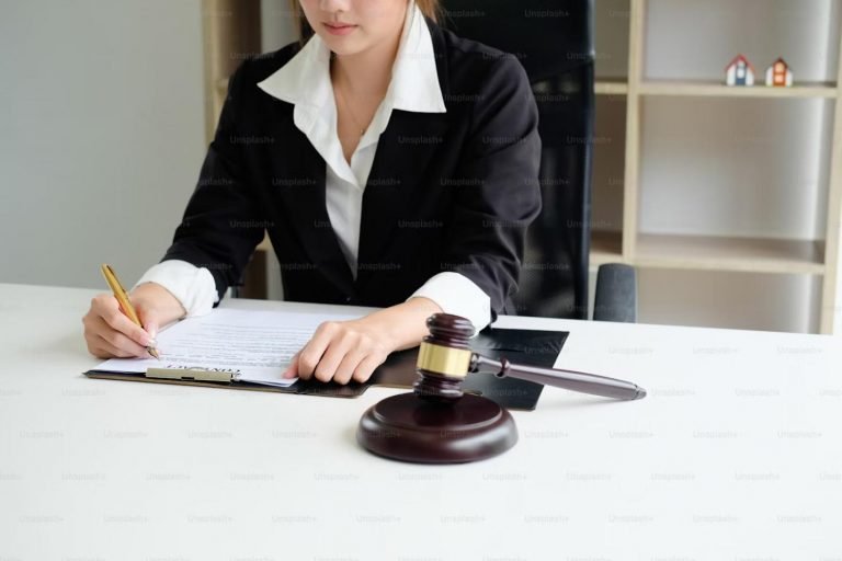 The image depicts a woman dressed in a black suit with a white collar, seated at a desk in an office setting.