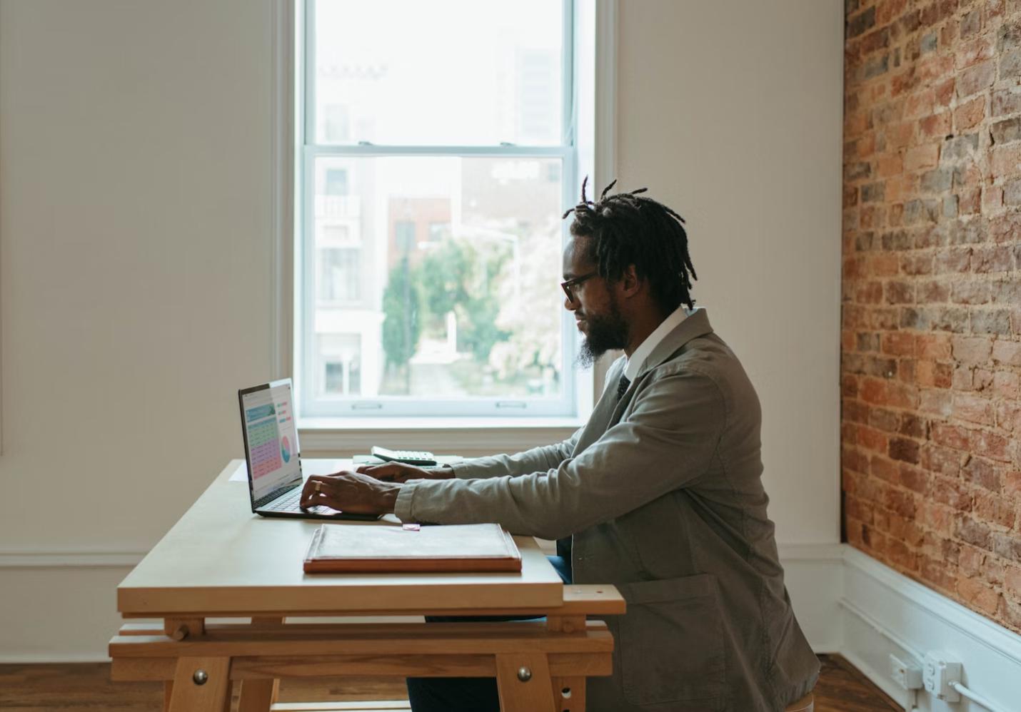 The image shows a man with dreadlocks and glasses, dressed in a casual yet professional attire, sitting at a wooden desk in a bright room with a large window