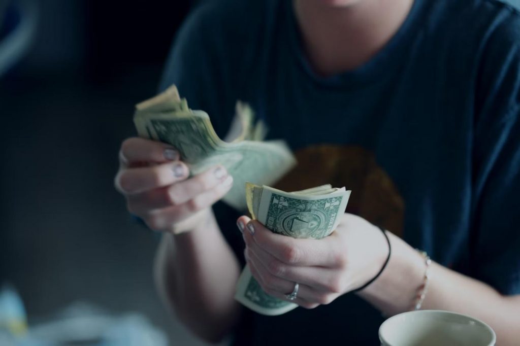 The image shows a close-up of a person’s hands as they count a stack of one-dollar bills. The person is wearing a casual t-shirt, and only the lower part of their face is visible.