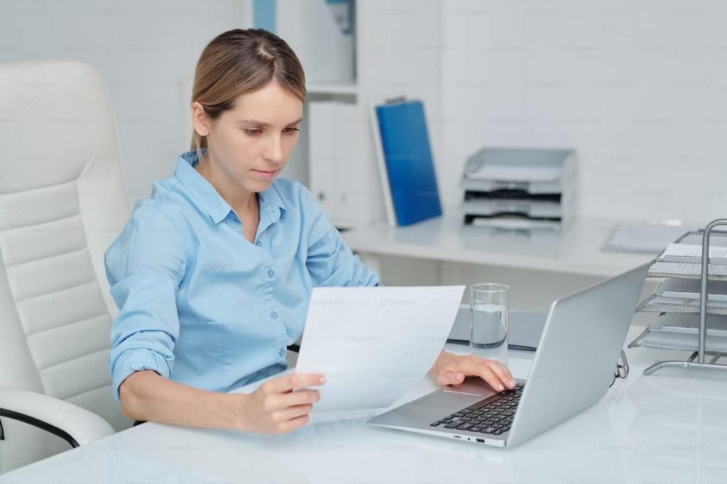 The image shows a woman in a light blue shirt sitting in a modern office environment. She is focused on reading a document she holds in her left hand while typing on a laptop with her right hand. 